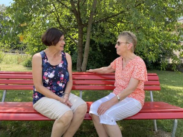 Bild: zwei Frauen des Besuchsdienstes sitzen auf einer roten Bank und hören einander zu.